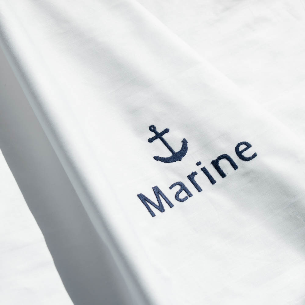 Dettaglio di lenzuolo bianco con ricamo blu di un'ancora e della scritta Marine
